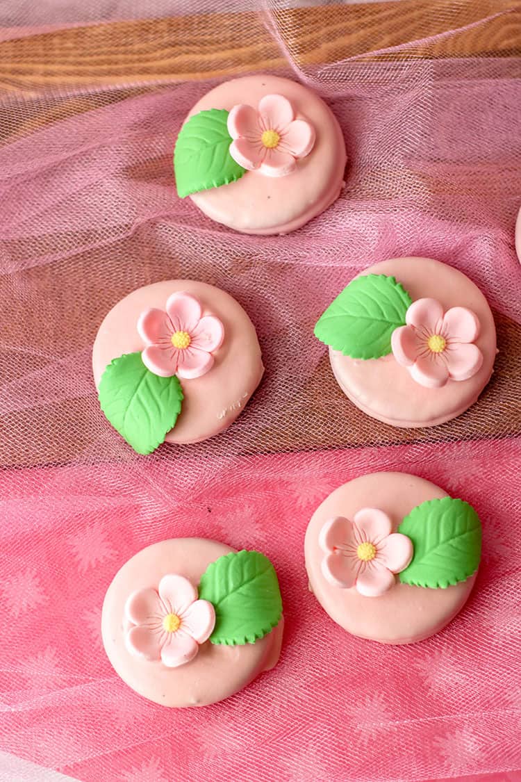 Five round pink biscuits decorated with pink flower and green leaf designs are arranged on pink and brown fabric.