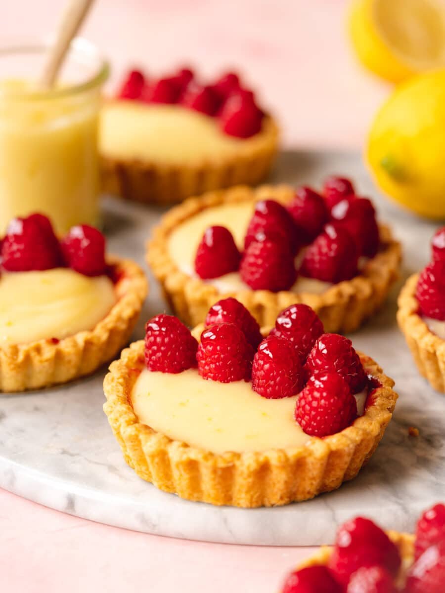 Small lemon tartlets with creamy filling topped with fresh raspberries are arranged on a marble surface, with lemons and a jar of curd in the background.