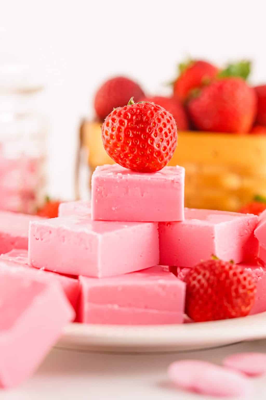 A stack of pink strawberry fudge squares is topped with a fresh strawberry, with more strawberries and fudge pieces in the background.