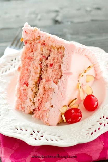 A slice of pink cherry almond cake with pink icing, garnished with cherries and almond flakes, on a white plate.