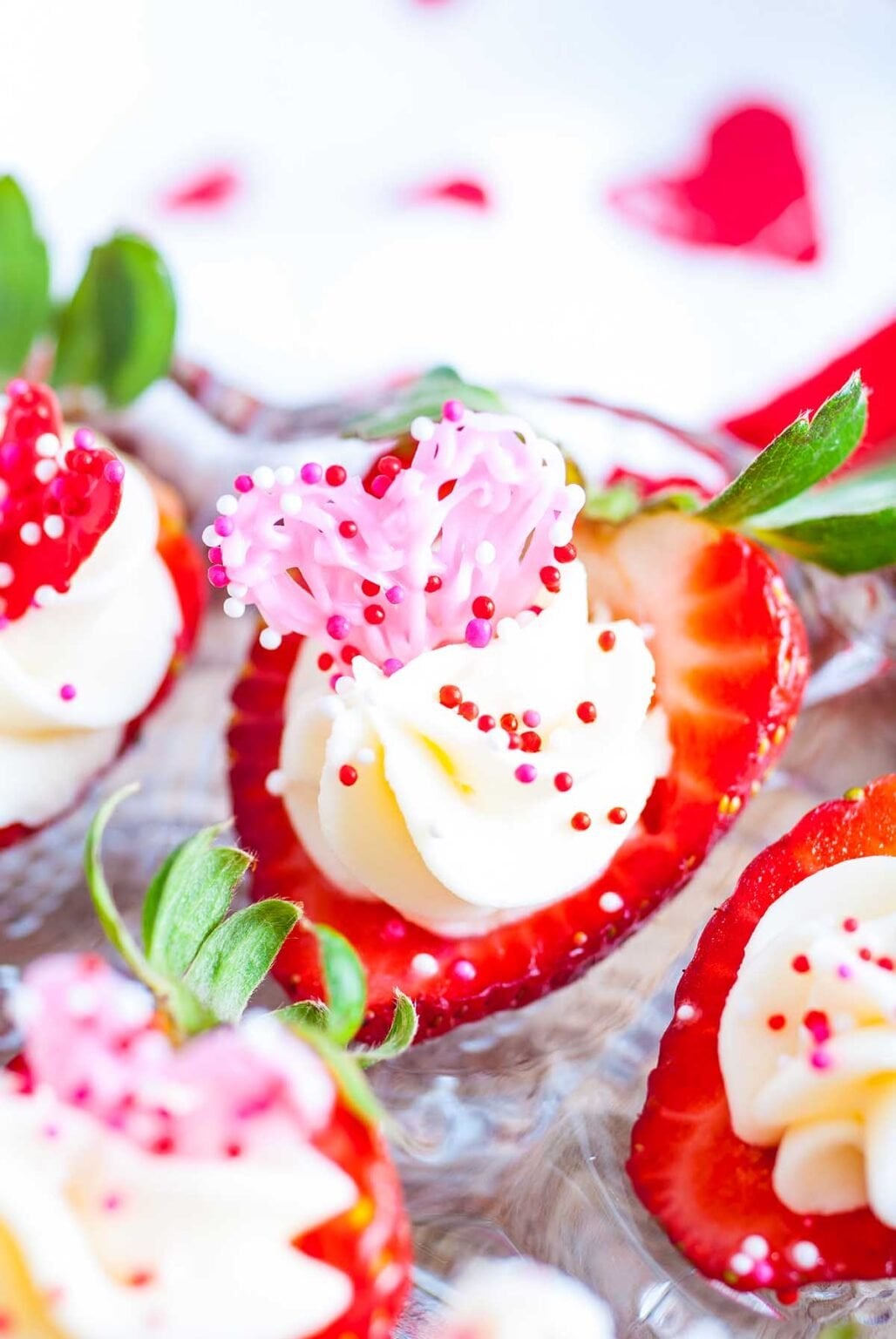Halved strawberries filled with swirls of white cream, topped with colourful pink and red sprinkles, displayed on a clear glass dish.