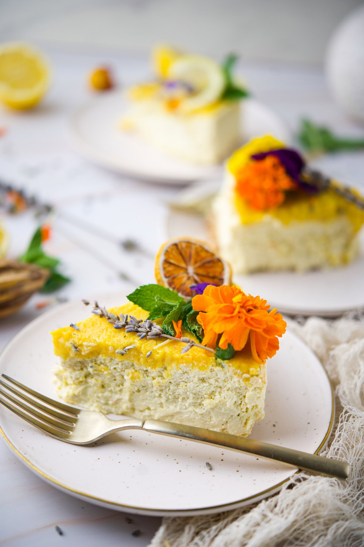 A slice of cake topped with yellow icing, edible flowers, and dried citrus sits on a white plate with a fork, with two more decorated cake slices in the background.