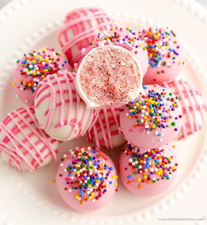 A plate of pink and white cake balls, some topped with rainbow sprinkles and others drizzled with pink icing; one cake ball is cut open to show the inside.