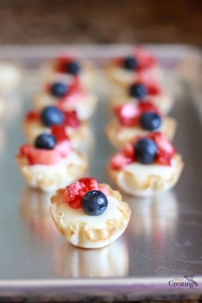 Mini tart cases filled with cream and topped with blueberries and diced strawberries, arranged on a metal baking tray.