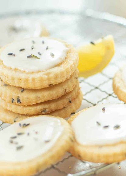 Stack of round, iced biscuits topped with lavender buds, arranged on a cooling rack with a lemon wedge in the background.