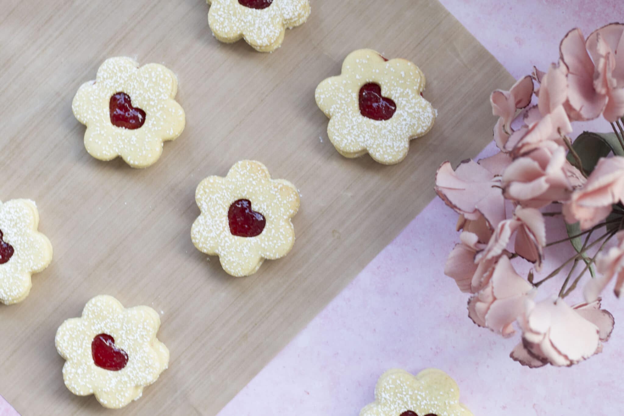 Flower-shaped biscuits with icing sugar and red jam centres are arranged on a light wooden surface, next to pink flowers on a pink background-perfect as cute Mother's Day desserts.