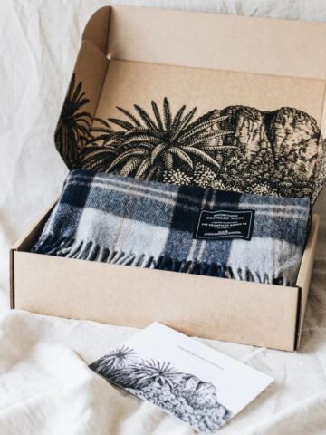 A blue and grey tartan blanket in an open cardboard box with black botanical illustrations; a matching postcard lies nearby on a light fabric surface, highlighting care instructions and fibre compression for easy storage.