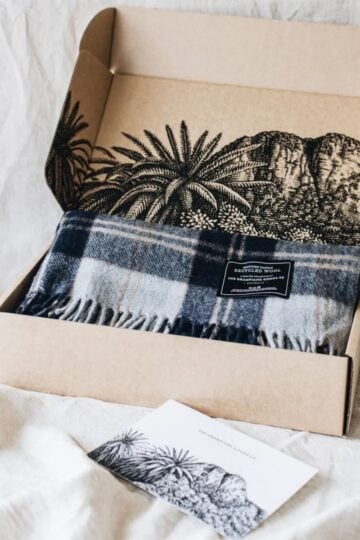 A blue and grey tartan blanket in an open cardboard box with black botanical illustrations; a matching postcard lies nearby on a light fabric surface, highlighting care instructions and fibre compression for easy storage.