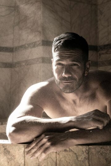 Shirtless man with short dark hair leans on the edge of a marble pool in a sunlit, tiled room, enjoying recovery equipment after an intense weekly training session.