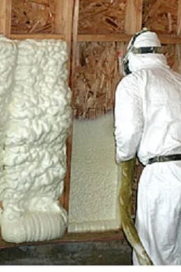 Person in protective gear spraying foam insulation material onto wooden wall studs inside a building under construction, ensuring the right insulation choice for optimal performance across different climate zones.