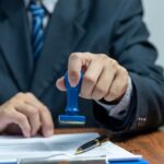 A person in a suit, possibly a migration agent, holds a blue stamp over a document on a desk, with a pen placed nearby, preparing for signing representation.