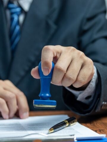A person in a suit, possibly a migration agent, holds a blue stamp over a document on a desk, with a pen placed nearby, preparing for signing representation.