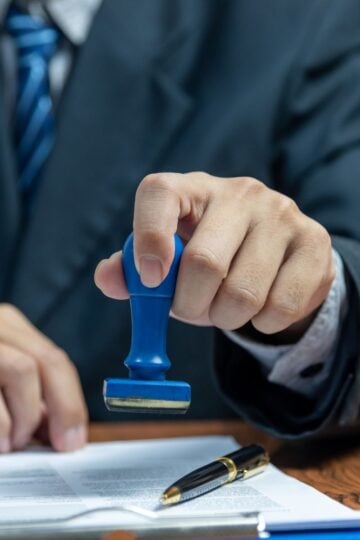 A person in a suit, possibly a migration agent, holds a blue stamp over a document on a desk, with a pen placed nearby, preparing for signing representation.