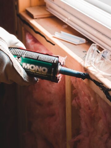 A gloved hand applies black sealant to a wooden window frame using a tube labelled
