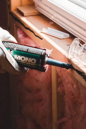 A gloved hand applies black sealant to a wooden window frame using a tube labelled “MONO,” making temporary repairs; safety spectacles rest nearby on the frame, ensuring work meets insurance standards.