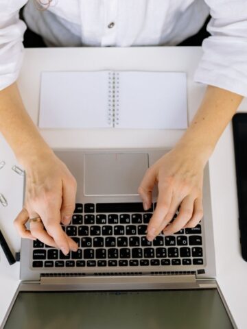 Person wearing a white shirt typing on a laptop at a desk with a notebook, pen, paper clips, and a tablet.