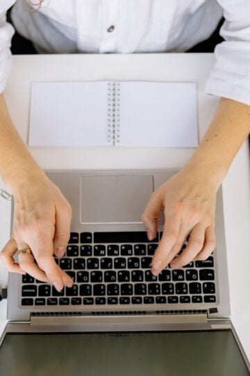 Person wearing a white shirt typing on a laptop at a desk with a notebook, pen, paper clips, and a tablet.
