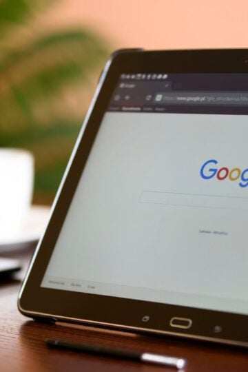 A Samsung tablet on a wooden table displays the Google homepage in a web browser, with a stylus and a blurred coffee cup in the background.