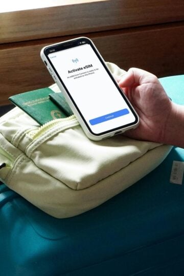 A person holds a mobile phone showing an “Activate eSIM” screen above a teal suitcase, ready for family life adventures or travels to the UK, with a passport and small bag resting on top.