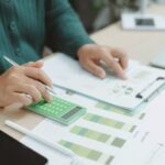 Person using a green calculator and examining financial charts and graphs on a desk with documents and a pen.