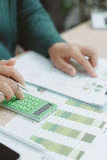 Person using a green calculator and examining financial charts and graphs on a desk with documents and a pen.
