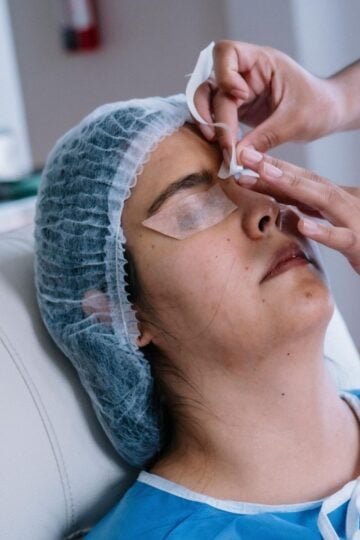 A person with a hair net and protective covering lies back as another applies a strip to their eyebrow, possibly for a cosmetic or medical procedure at Becker Plastic Surgery, aiming for natural-looking results.