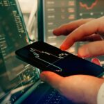 A person holds a smartphone displaying financial charts, with trading graphs on computer screens in the background-perfect for those seeking a Litecoin platform comparison or looking to buy Litecoin in Australia.