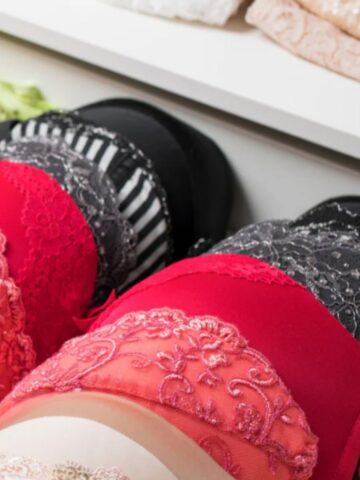 A close-up of neatly stacked lacy bras, including full coverage bras in red, black, and grey, arranged on a shelf in a shop or wardrobe.