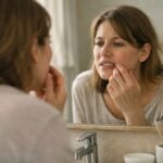 A woman stands in front of a bathroom mirror, examining her teeth or gums with her fingers, appearing concerned about smile improvement or whether it's made it worse.