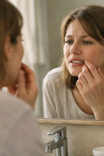 A woman stands in front of a bathroom mirror, examining her teeth or gums with her fingers, appearing concerned about smile improvement or whether it’s made it worse.