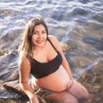A pregnant woman in maternity swimwear kneels in shallow water by the shore, smiling at the camera.