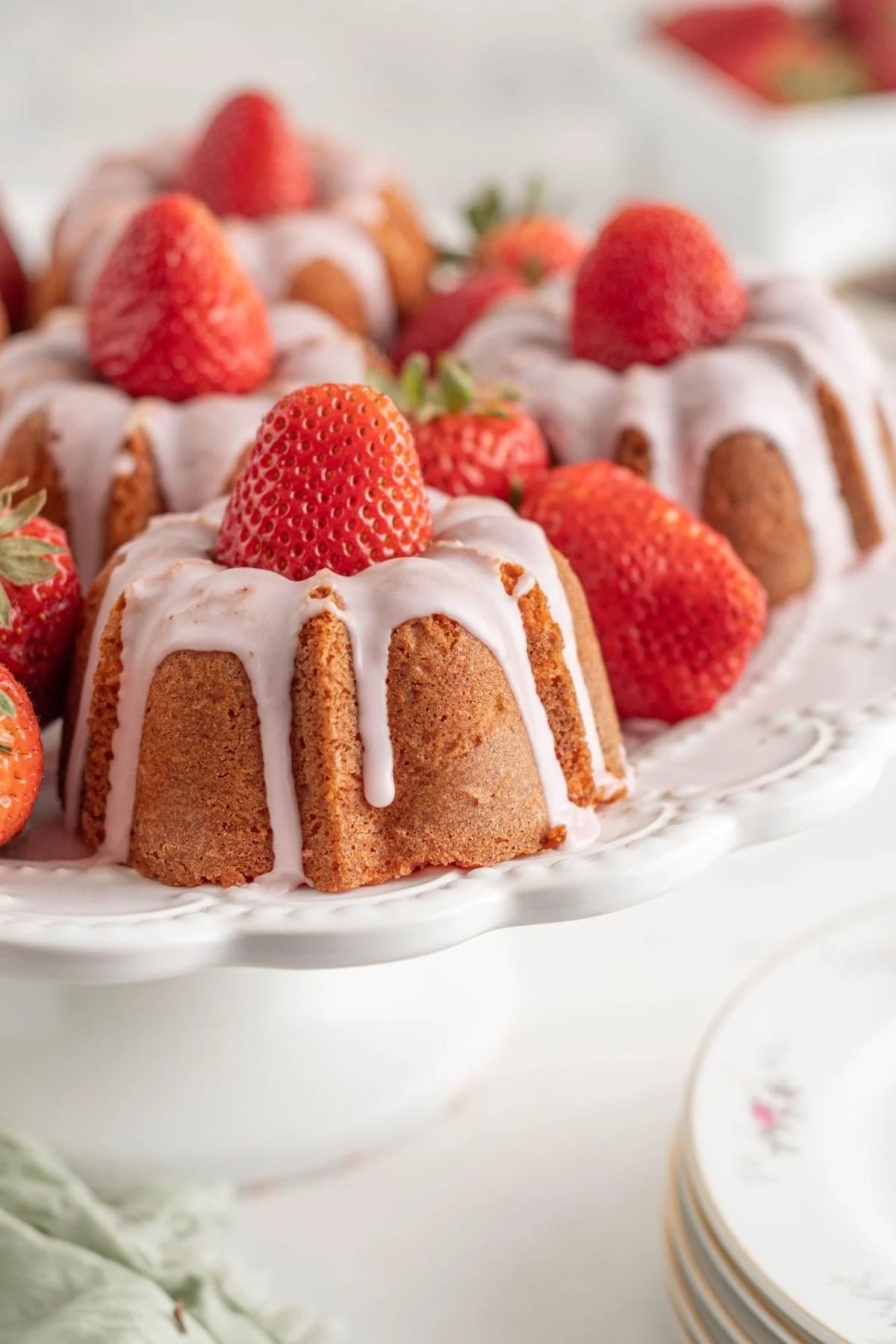 Mini bundt cakes topped with white icing and whole strawberries are arranged on a white cake stand, with more strawberries in the background.