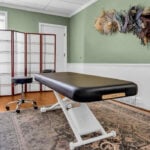 A massage table in a well-lit room with green walls, a decorative rug, potted plant, folding screen, and wall-mounted dried floral arrangement creates the perfect spa oasis for those looking to build a luxury spa experience at home.