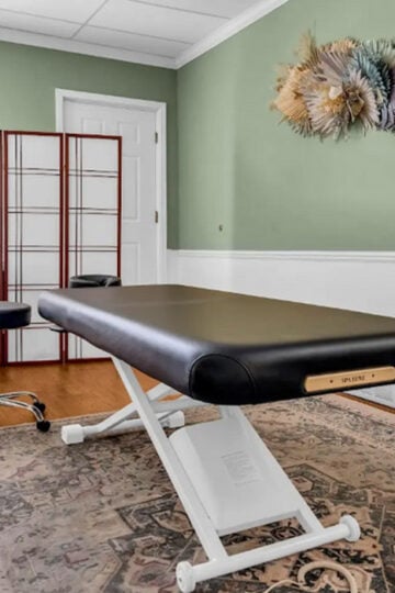 A massage table in a well-lit room with green walls, a decorative rug, potted plant, folding screen, and wall-mounted dried floral arrangement creates the perfect spa oasis for those looking to build a luxury spa experience at home.