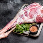 Organic Meat UK: Raw lamb leg on a metal tray with a small bowl of pink Himalayan salt and fresh mint leaves, set on a dark surface, showcasing premium organic meat quality.