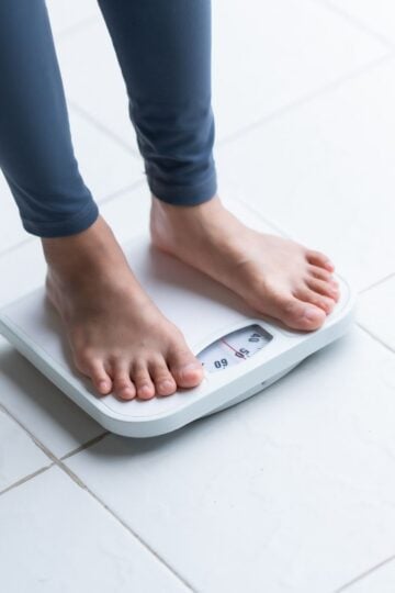 A person standing barefoot on a white bathroom scale, checking their weight on a tiled floor—a common scene for anyone tracking progress during a weight loss journey or exploring new options like Cagrilintide and other weight loss peptides.