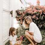 An adult and a child, both in white shirts, are crouching by flowering shrubs in the garden near a white building, enjoying family life on a sunny day.