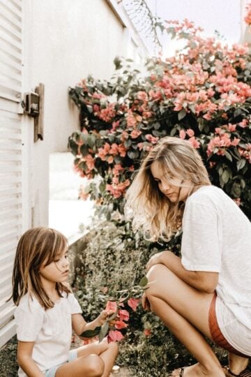 An adult and a child, both in white shirts, are crouching by flowering shrubs in the garden near a white building, enjoying family life on a sunny day.
