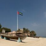 A large UAE flag on a tall pole stands behind an old wooden boat and palm trees on a sandy beach under a clear sky.