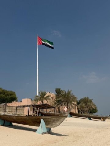 A large UAE flag on a tall pole stands behind an old wooden boat and palm trees on a sandy beach under a clear sky.