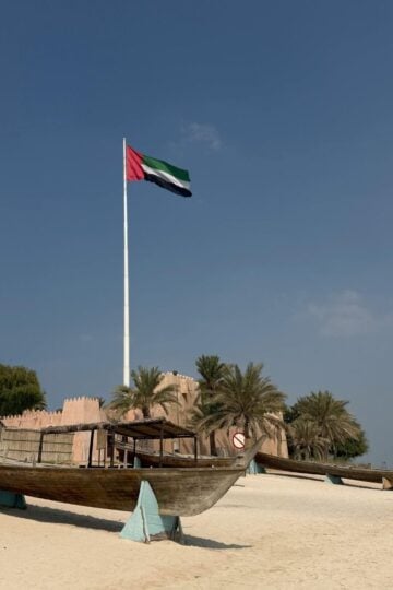 A large UAE flag on a tall pole stands behind an old wooden boat and palm trees on a sandy beach under a clear sky.