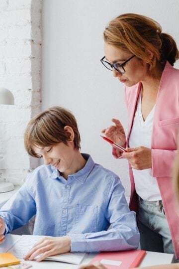 A teacher in a pink blazer stands beside a smiling pupil who is drawing at a desk, surrounded by plants and school supplies that reflect a balanced lifestyle.