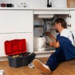 A skilled plumber in blue overalls repairs pipes under a kitchen sink in San Jose, with an open toolbox containing tools on the floor nearby.
