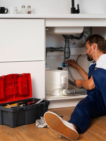 A skilled plumber in blue overalls repairs pipes under a kitchen sink in San Jose, with an open toolbox containing tools on the floor nearby.