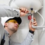 A reliable plumber from Milpitas, wearing safety glasses and a cap, uses a spanner to work on pipes and valves underneath a sink or water heater-ensuring quality home maintenance.
