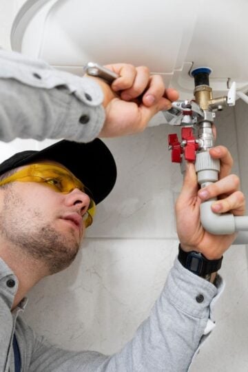 A reliable plumber from Milpitas, wearing safety glasses and a cap, uses a spanner to work on pipes and valves underneath a sink or water heater—ensuring quality home maintenance.