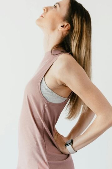 Woman in sportswear stands with hands on lower back, head tilted back as if stretching to relieve discomfort, highlighting the importance of skin elasticity and healthy breast tissue against a plain white background.