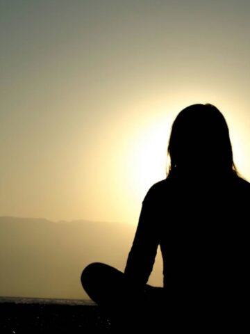 A person sits cross-legged outdoors, silhouetted against a sunset or sunrise with mountains in the background, embracing a moment of self-discovery.