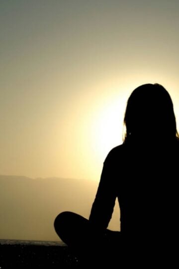 A person sits cross-legged outdoors, silhouetted against a sunset or sunrise with mountains in the background, embracing a moment of self-discovery.