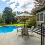 A garden with a round swimming pool featuring warmer pools for comfort, a concrete patio, table with four chairs and a large parasol, next to a house with steps, surrounded by trees and greenery.